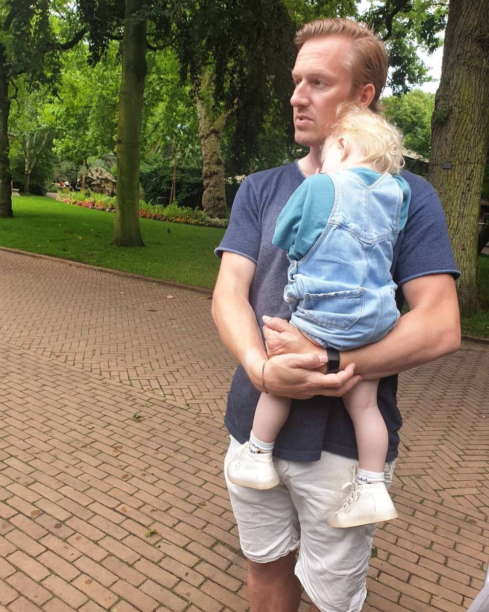 A man carrying a toddler on his back on a paved path through a park.