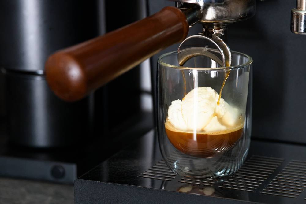 A double-walled glass filled with a ball of vanilla ice cream sitting under a coffee machine, getting poured over with an espresso.