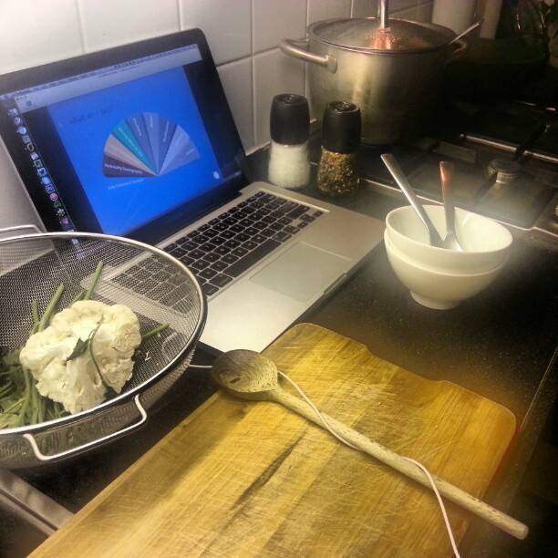 A laptop displaying a color chart sits on a kitchen counter next to a colander of cauliflower and a pot on the stove.