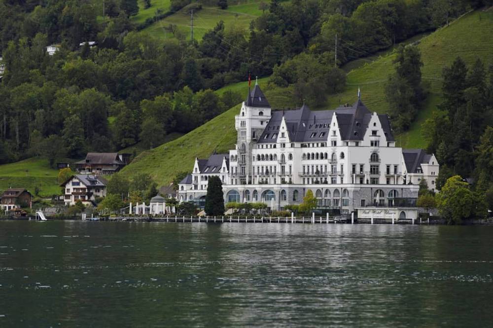 A white castle-like hotel building stands beside a lake with a green hillside behind it.