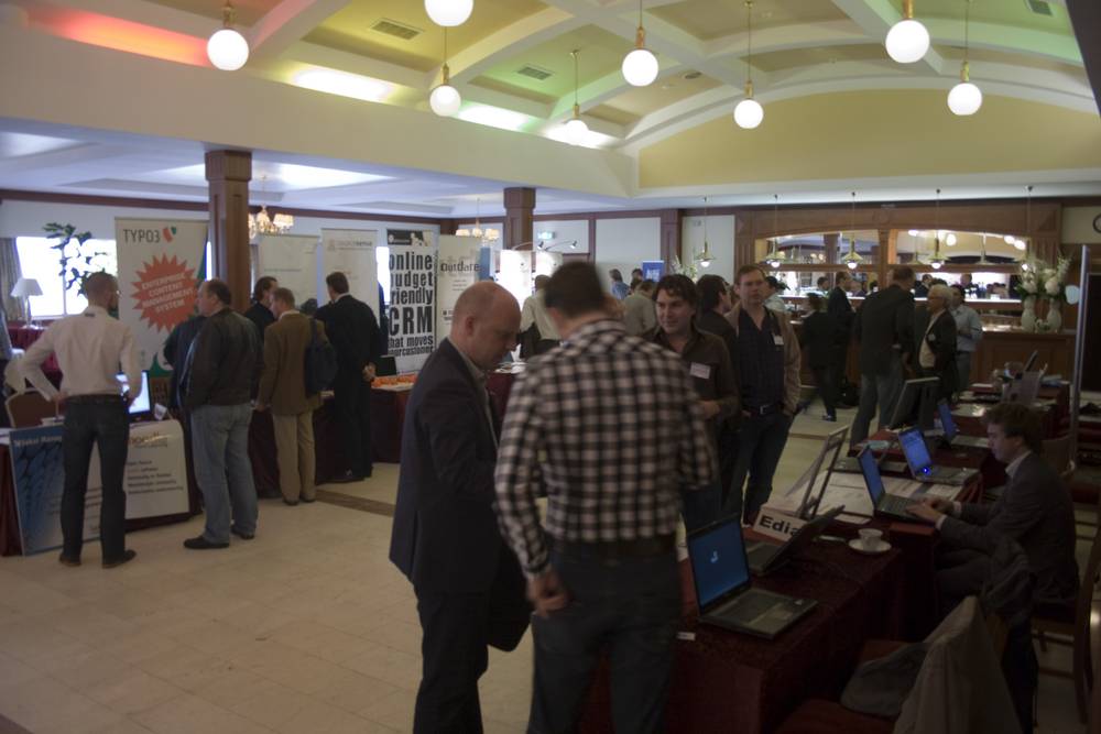 Conference attendees browsing exhibitor booths and laptops in a large hall with vaulted ceilings.