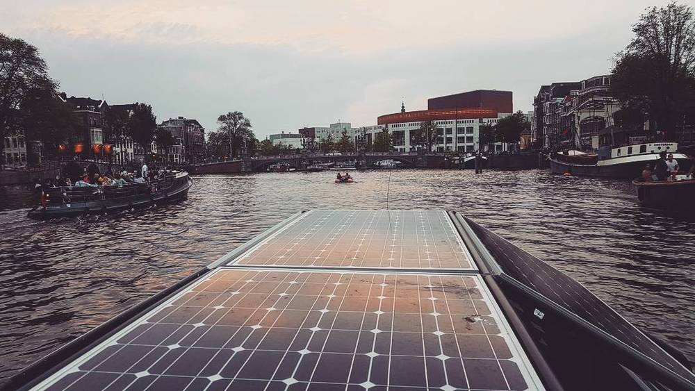 A solar-panelled boat gliding along an Amsterdam canal lined with houseboats and buildings.