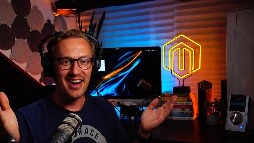 A man wearing headphones and gesturing while recording at a desk, with a glowing orange hexagonal logo lit up behind him.
