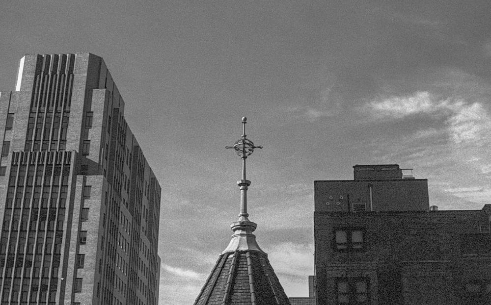 View across rooftops toward a slender metal spire crowned with an ornate cross, rising from a small conical roof. To the left, a tall mid-century tower with vertical concrete fins fills the frame; to the right, the dark brick upper stories of another building with mechanical penthouses. Wispy clouds streak across the sky behind the cross.