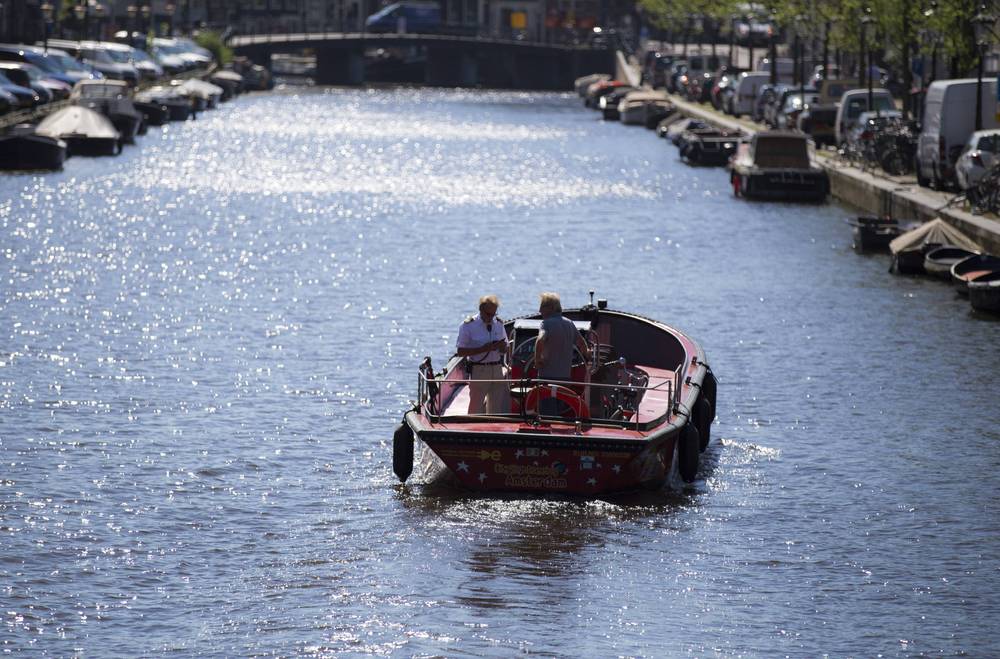 A small boat carries passengers down a sunlit Amsterdam canal lined with houseboats.