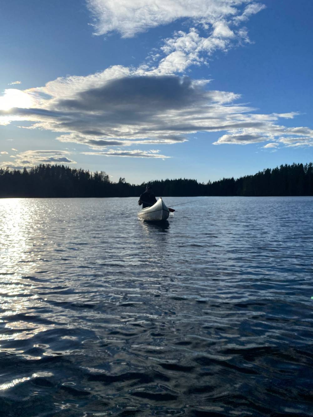 A lone figure fishing from a white canoe on a calm lake, silhouetted against a wide sky with dramatic cloud formations. Dense conifer forest lines the horizon. The low sun reflects off the rippling water in the foreground.