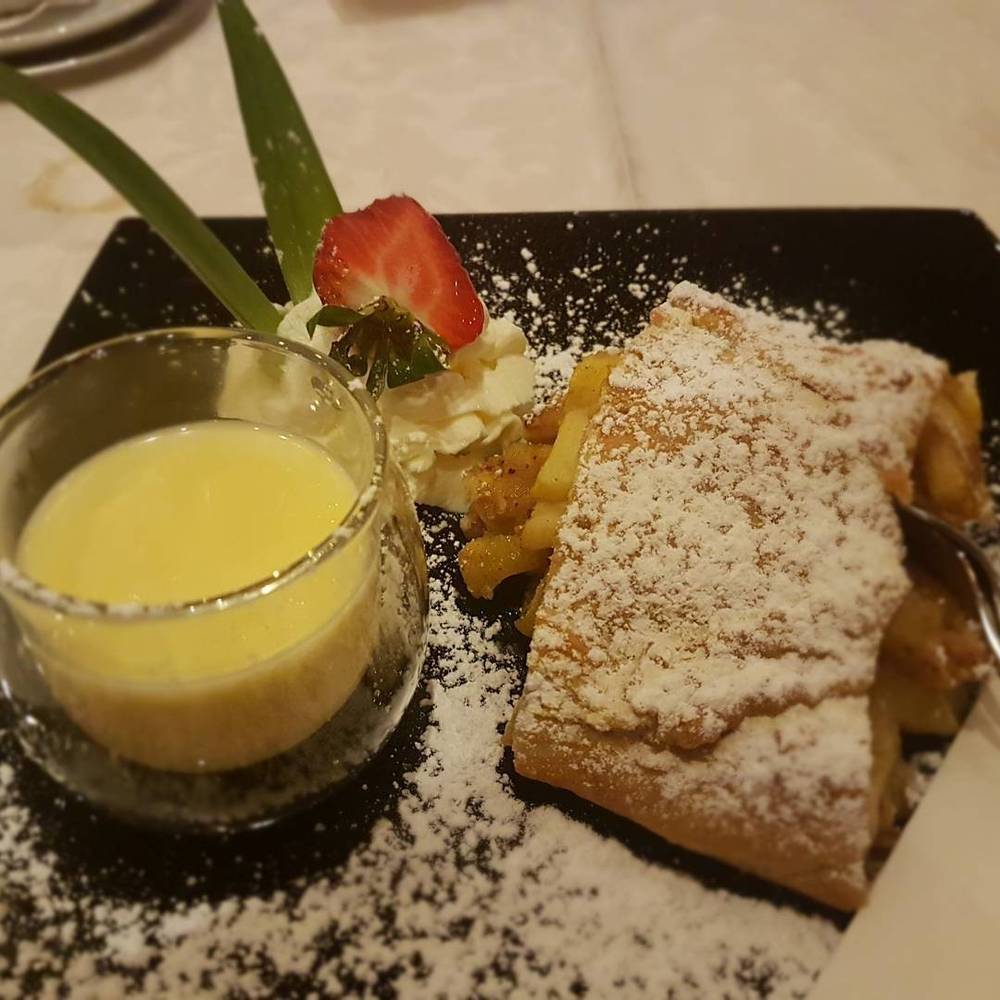 An apple strudel dessert served with vanilla sauce, garnished with a strawberry.
