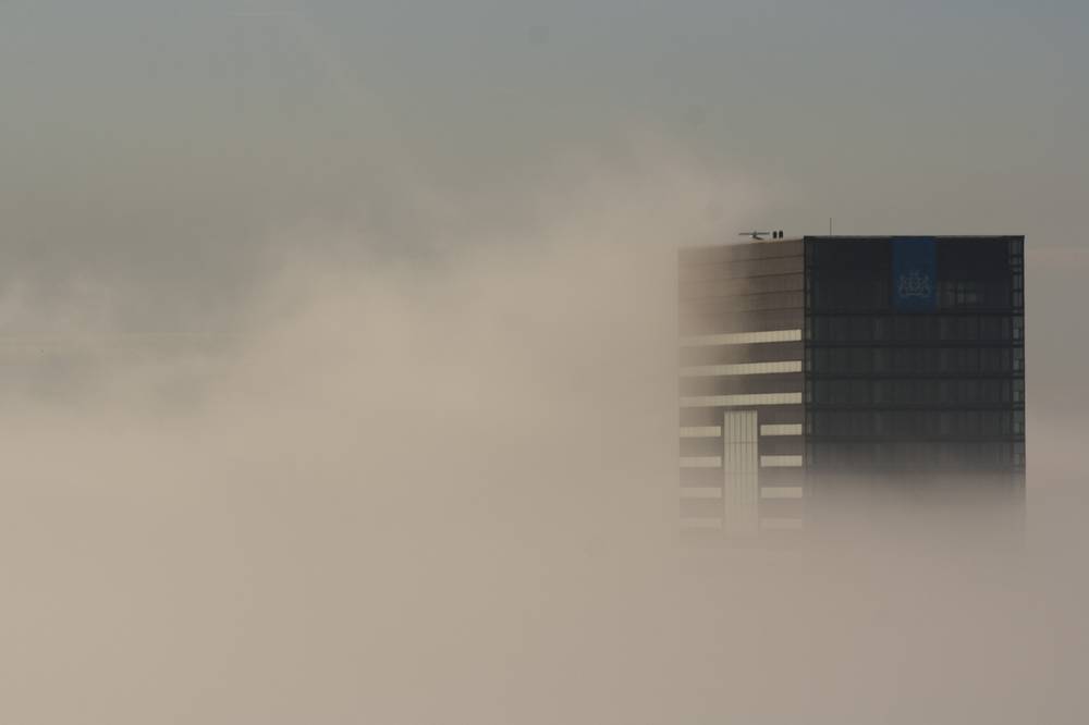 A high-rise building emerges from dense fog, its lower floors swallowed by the mist.