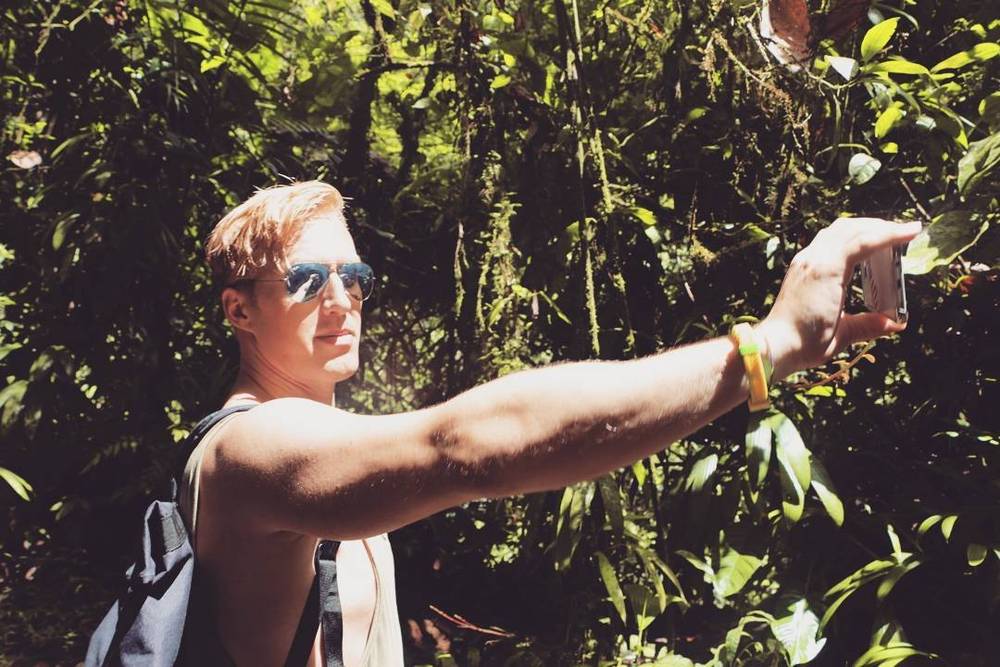 A man taking a photo with his phone while standing among green foliage.