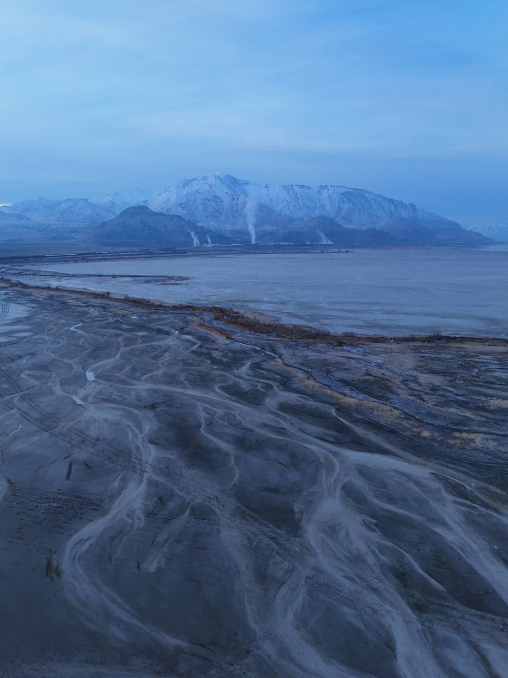 The foreground is dominated by a vast, textured expanse of frozen mud or silt, marked by intricate, vein-like patterns of ice and dried earth that curve and branch across the surface like a dried riverbed. Beyond this textured ground lies a wide, flat body of frozen water or ice sheet that stretches horizontally across the frame, bordered by a thin strip of brown, dormant vegetation along the shoreline. In the distance, a majestic range of snow-capped mountains rises against the sky, their peaks partially obscured by a light haze or low clouds, creating a layered backdrop. At the base of the mountains, faint plumes of white steam or smoke rise vertically from what appears to be an industrial facility, contrasting with the natural landscape. The entire scene is bathed in the cool, monochromatic blue light of twilight or early dawn, creating a soft, diffuse illumination with no harsh shadows. The viewpoint is elevated, likely from a drone or high vantage point, looking down at an angle t