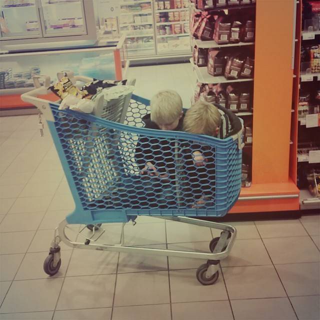 A shopping cart with two children seated in the child seat, parked in a supermarket aisle.