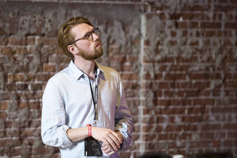 A bearded man in glasses stands thoughtfully against a brick wall at the conference.