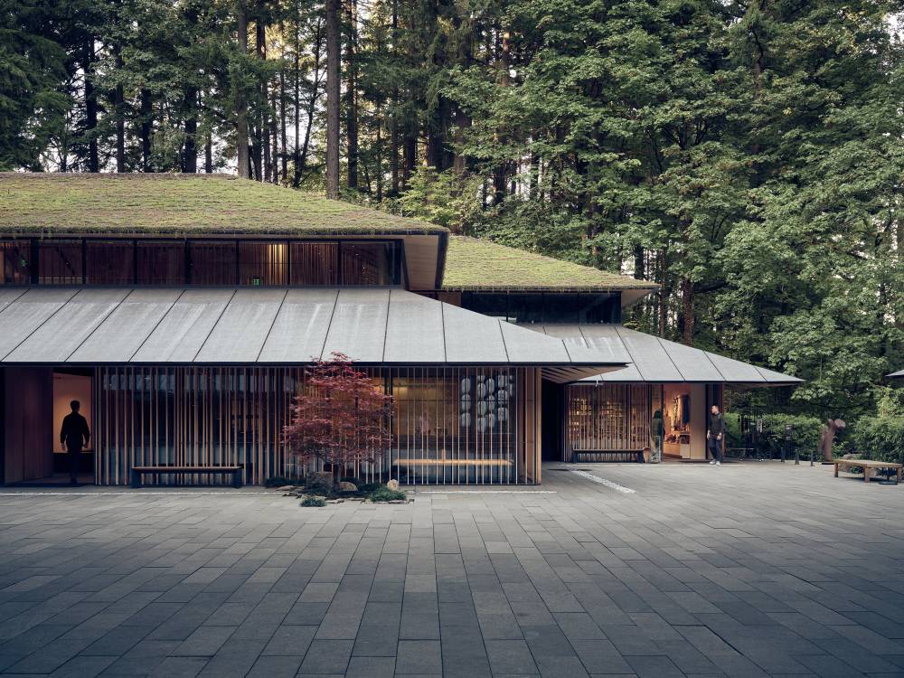 A striking contemporary pavilion emerges from the Portland Japanese Garden, its grass-covered living roof seamlessly blending into the surrounding Pacific Northwest forest. The building's modernist design features vertical wooden slats, metal roofing, and floor-to-ceiling windows that reveal warm interior lighting at dusk. A solitary figure silhouetted in the left entrance adds human scale to the architectural composition, while a crimson Japanese maple provides a delicate counterpoint to the structure's bold horizontal lines.