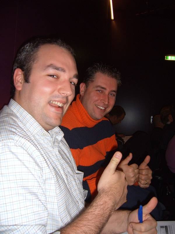 Two men grin and give thumbs up at an evening event.
