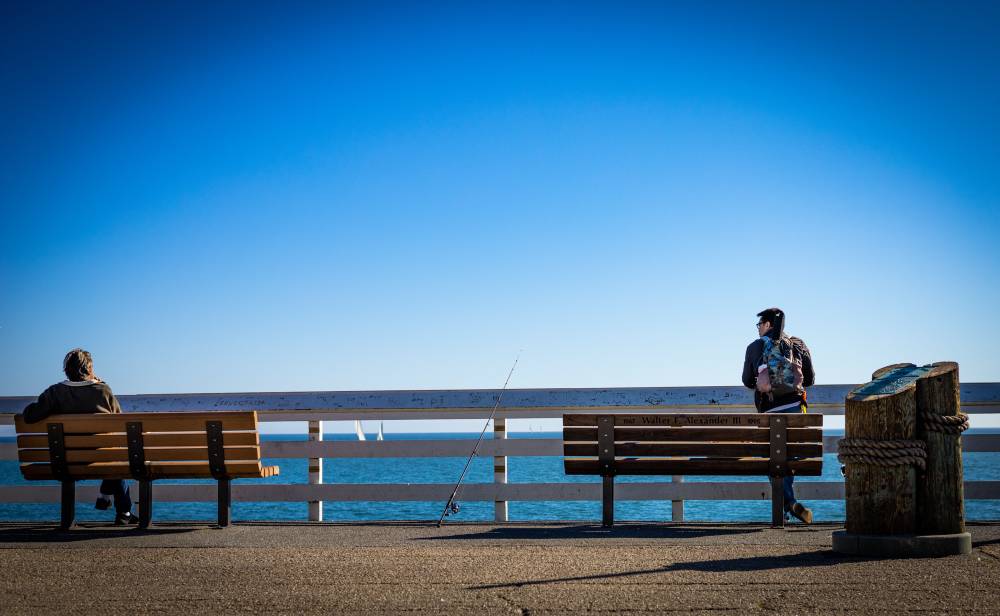A wide shot of a pier showing two people on opposite ends of two adjacent memorial benches, both facing the ocean. A fishing rod stands propped between them, and the expansive blue sky dominates the upper two-thirds of the frame.