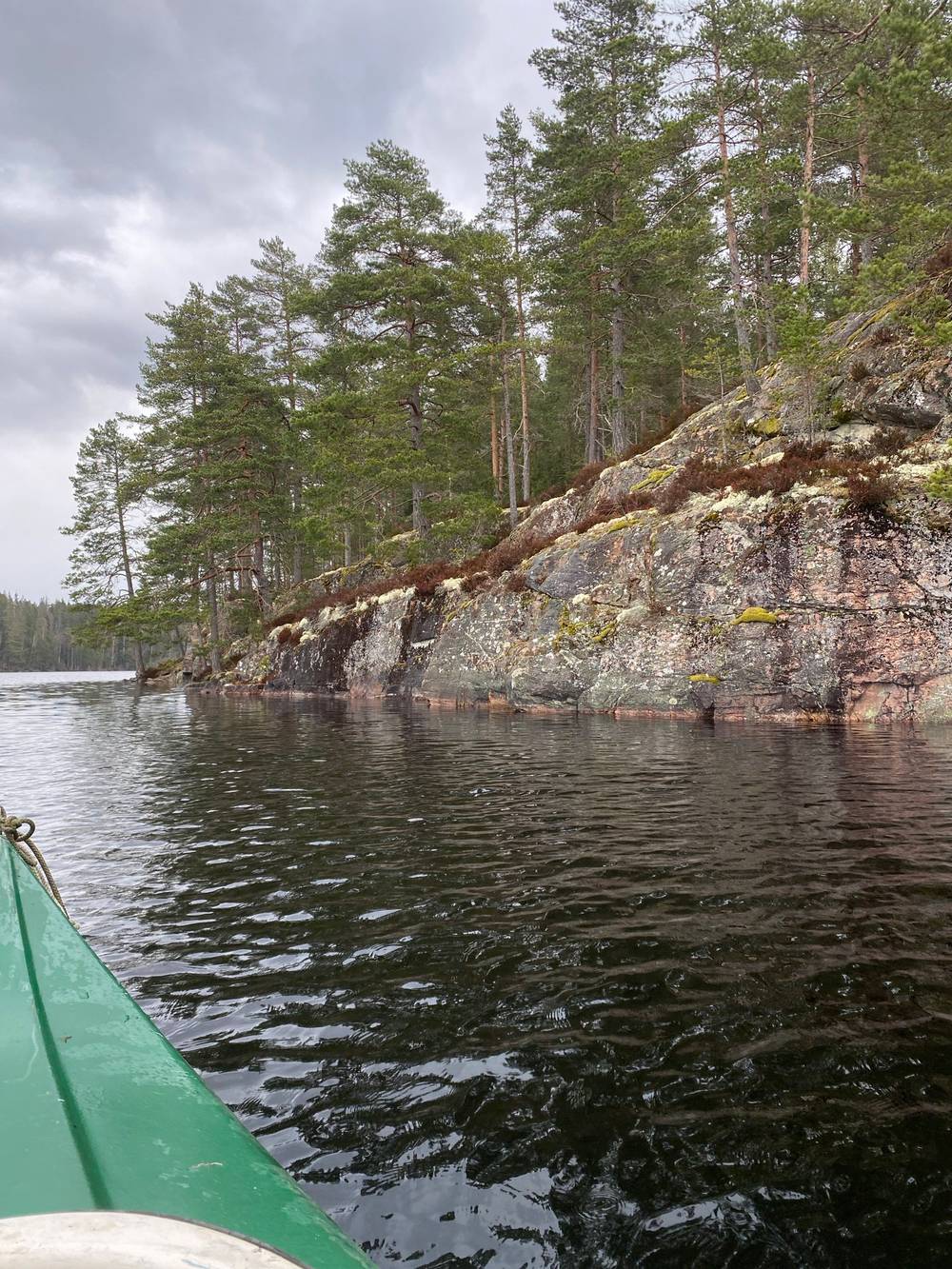 View from the bow of a green rowboat passing a lichen-covered granite cliff face topped with pine trees, under an overcast sky.