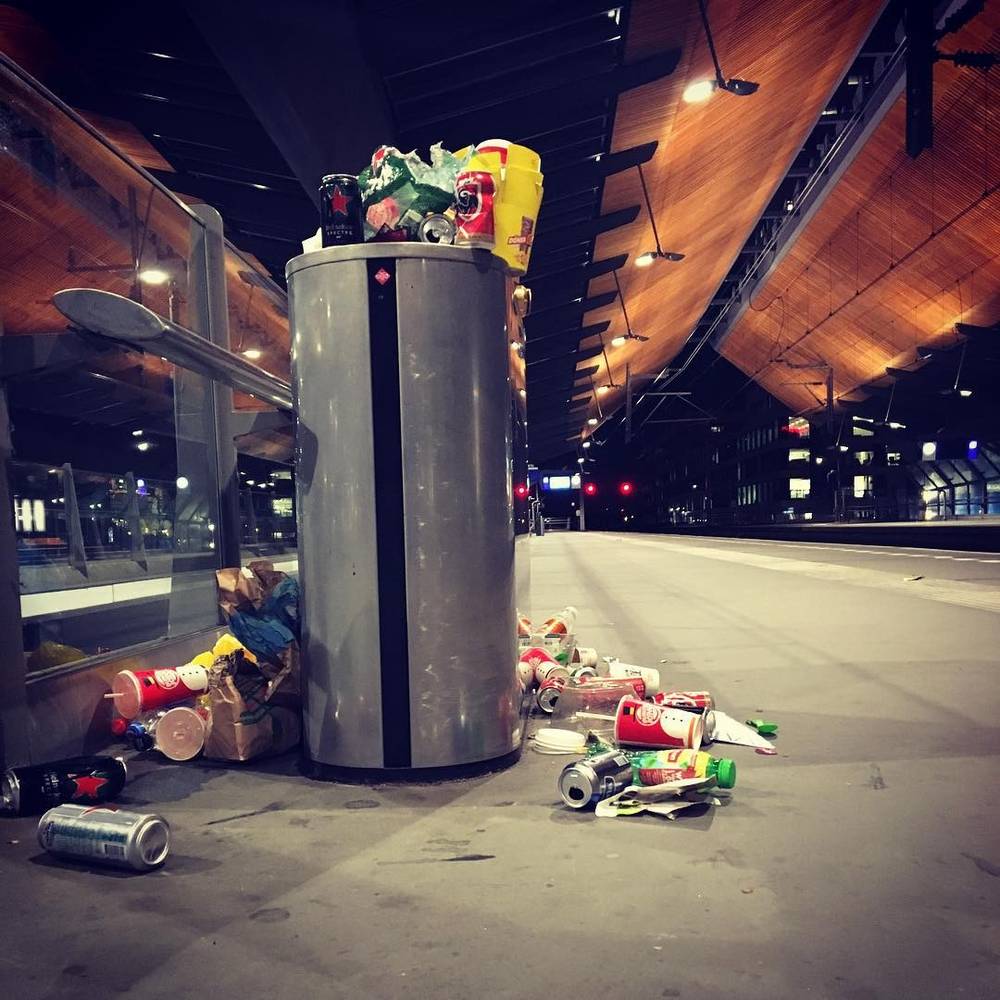 An overflowing trash can with cans and litter scattered around it in a train station.