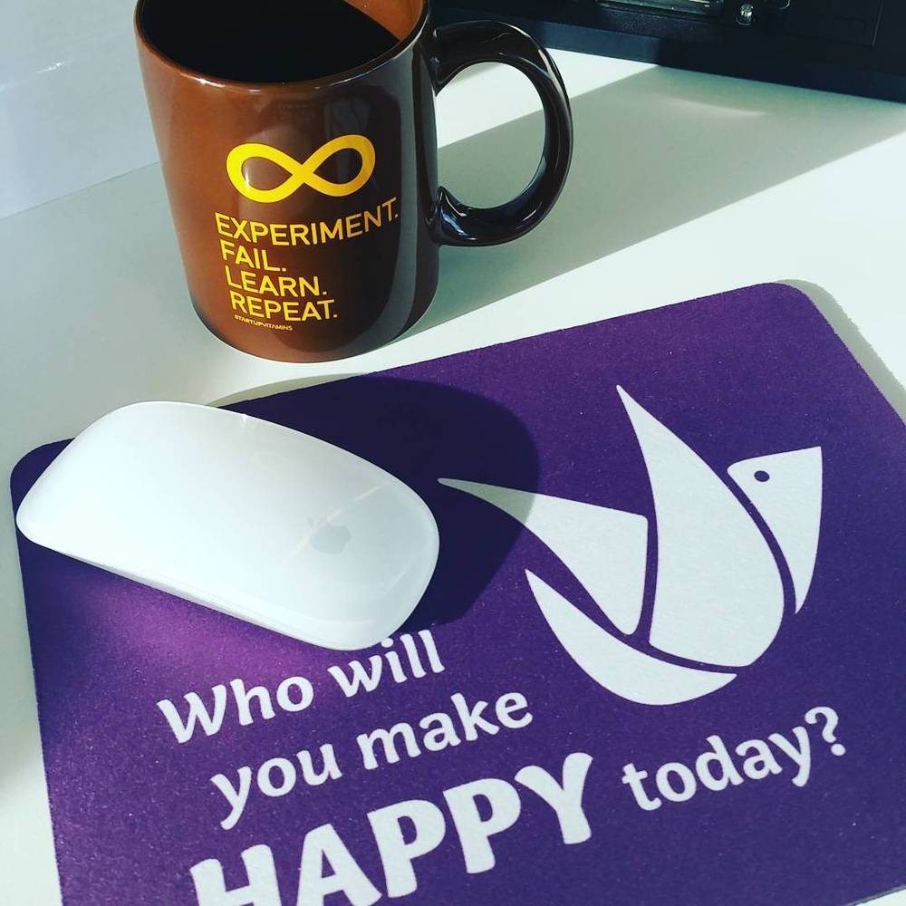 A black coffee mug reading "Experiment. Fail. Learn. Repeat." beside a mouse pad with the text "Who will you make happy today?".