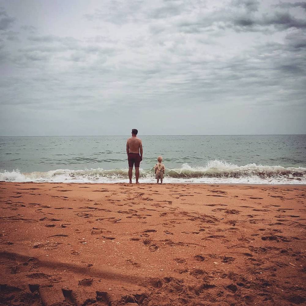 A man and a small child standing at the water's edge on a beach with reddish-brown sand.