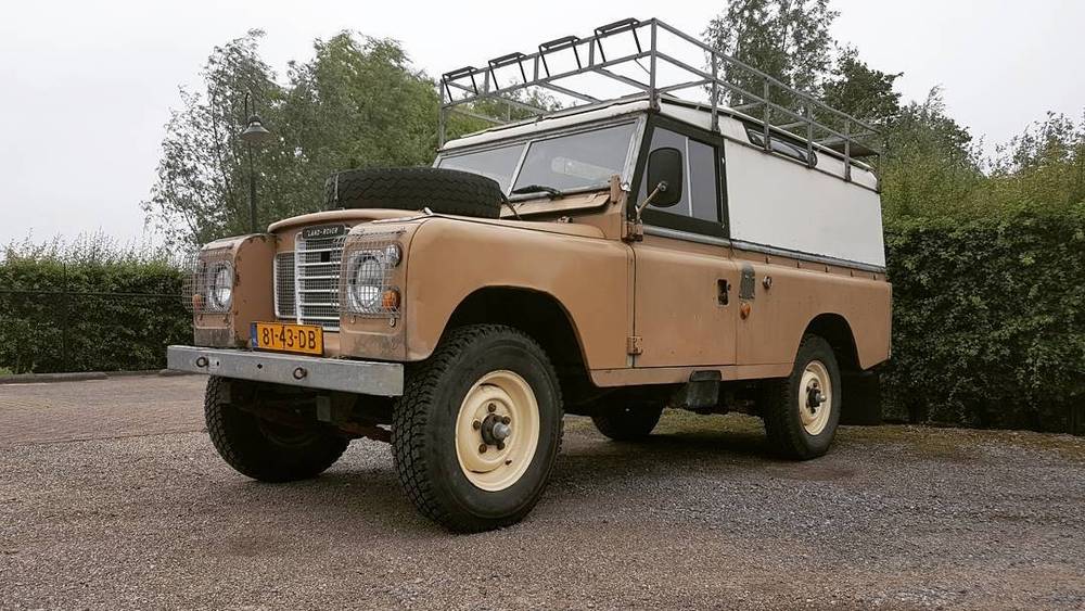 A restored beige Land Rover Series pickup with a roof rack, parked outdoors.