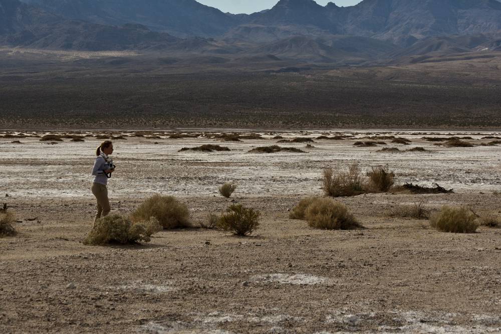 A woman stands alone photographing a vast, flat desert basin ringed by mountains.