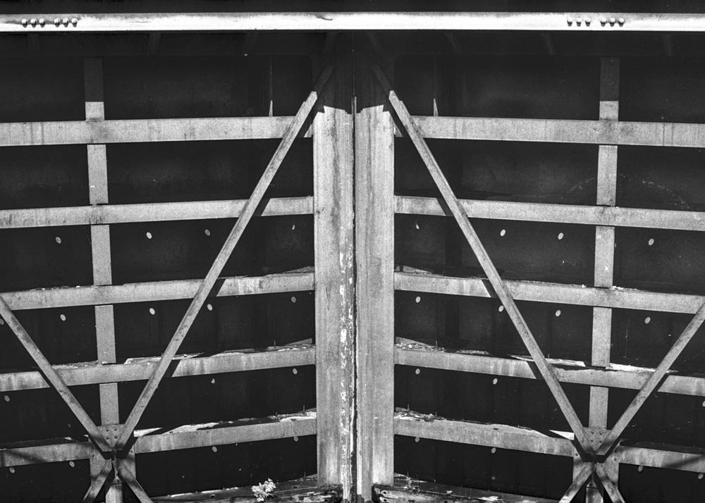Looking straight on at a pair of massive steel lock gates meeting in a tall central miter. Heavy horizontal girders stretch across each leaf, crossed by diagonal tension rods that draw an X through each panel. Bolt and rivet heads punctuate the bracing in a regular grid. The metal is streaked with darker water stains; below the gates, a glimpse of the lock chamber floor catches scattered light.