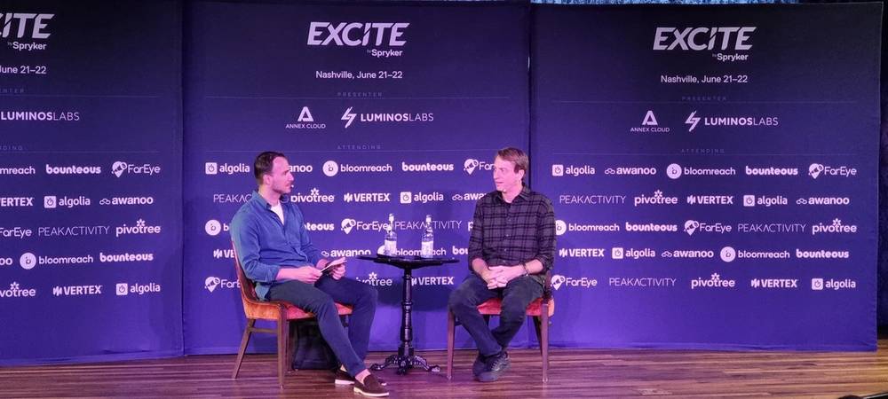 Two men seated in conversation on an Excite conference stage with sponsor logos displayed behind them.