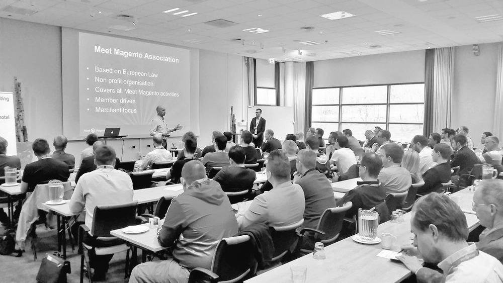 A black-and-white photo of a conference audience seated at tables listening to a talk.