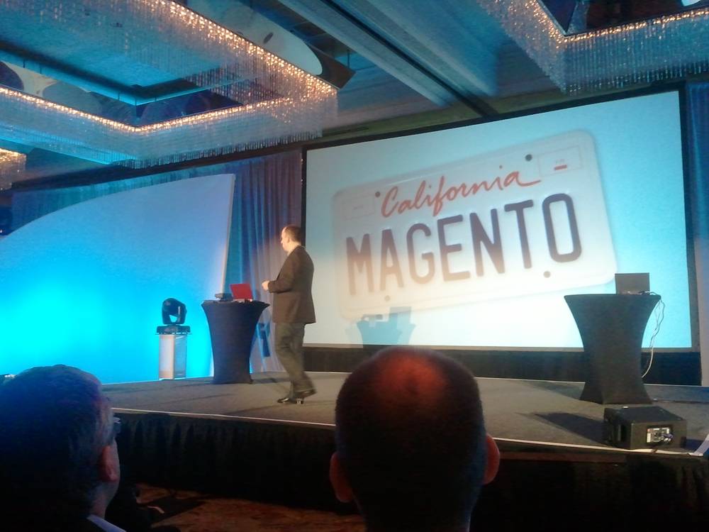 A presenter stands beside a screen showing a 'California Magento' license plate graphic.