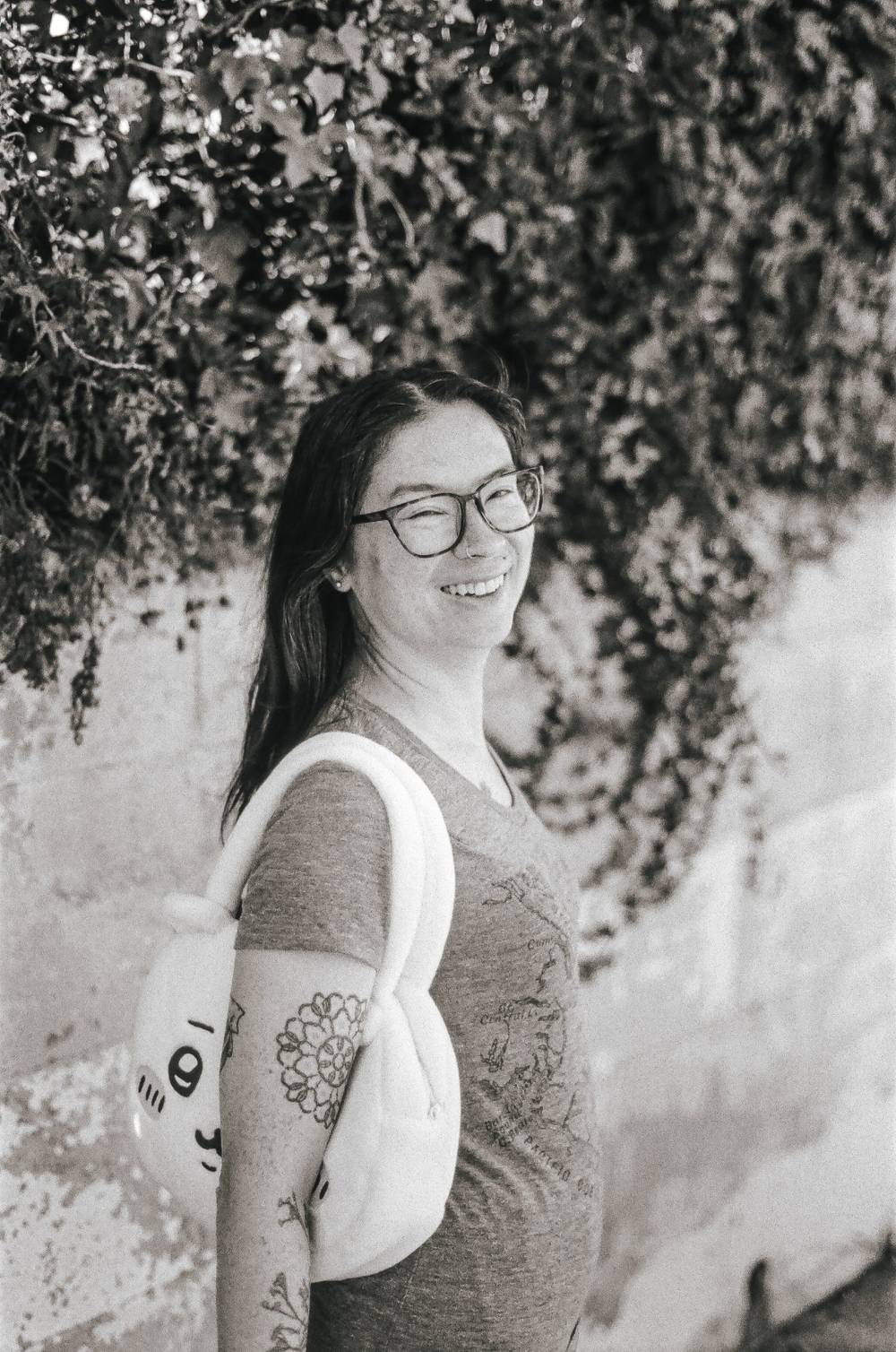 Erica. Black-and-white film portrait of a woman with long dark hair, glasses, and a floral arm tattoo, smiling over her shoulder while carrying a tote bag, standing in front of ivy-covered walls