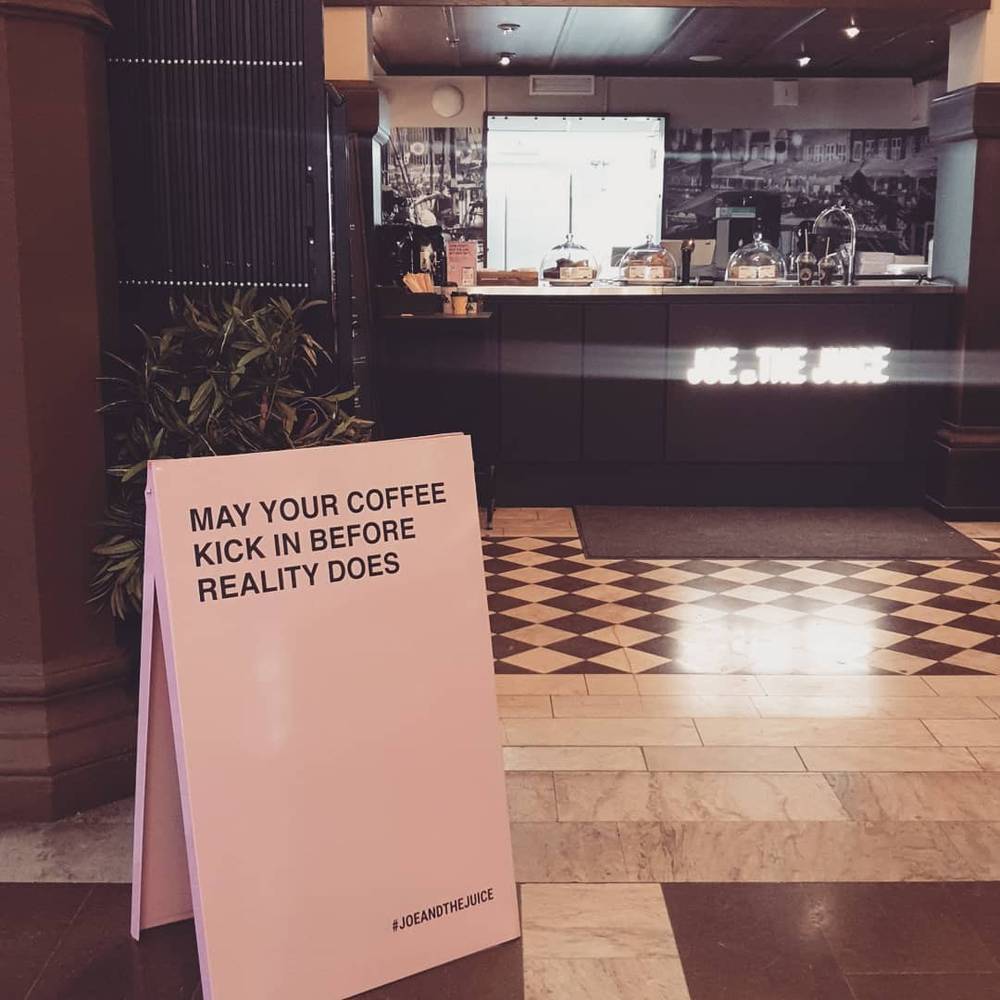 A sandwich board outside a Joe & The Juice shop reads "May your coffee kick in before reality does".