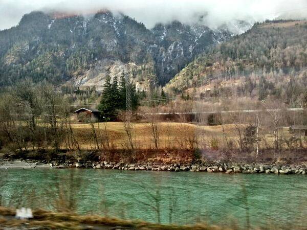A turquoise mountain river winds through a forested valley with snow-capped peaks in the background.