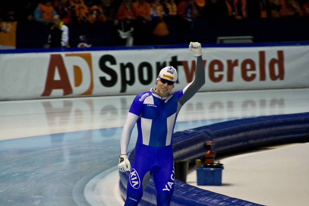 A speed skater in a blue suit raises a fist in celebration beside an "AD Sportwereld" banner.