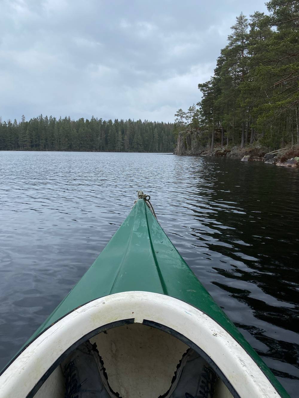 First-person view over the bow of a green rowboat crossing a dark, rippling lake, with a pine-forested shoreline and rocky outcrops ahead under cloudy skies.