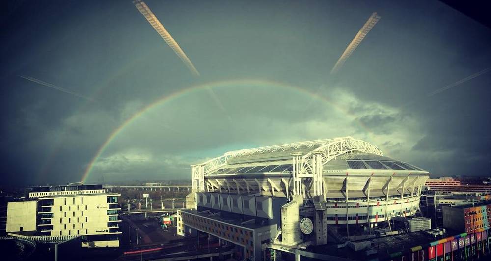A rainbow arching over a stadium and surrounding buildings under a stormy sky.