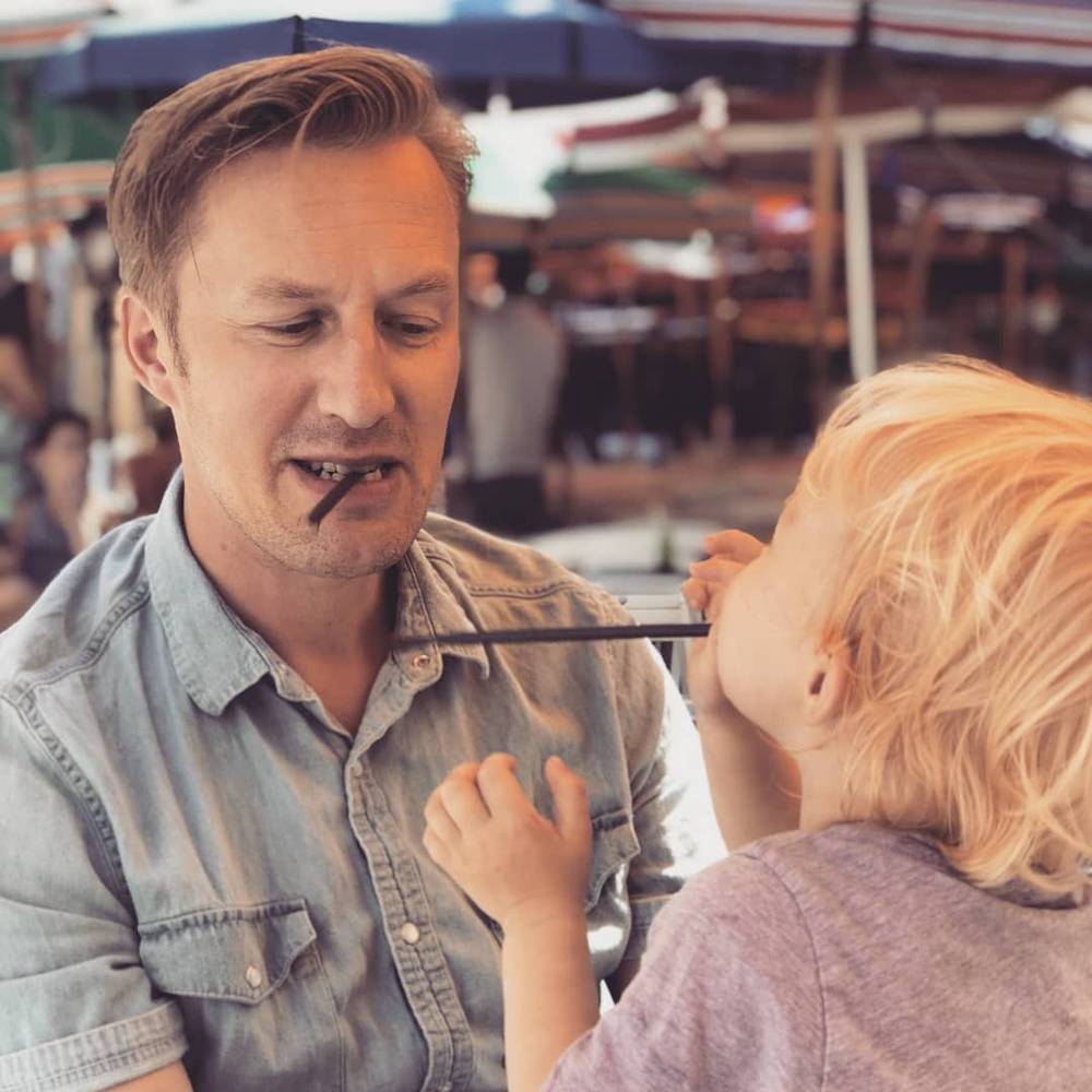 A man and a toddler playfully tug on opposite ends of a striped drinking straw held in their teeth.