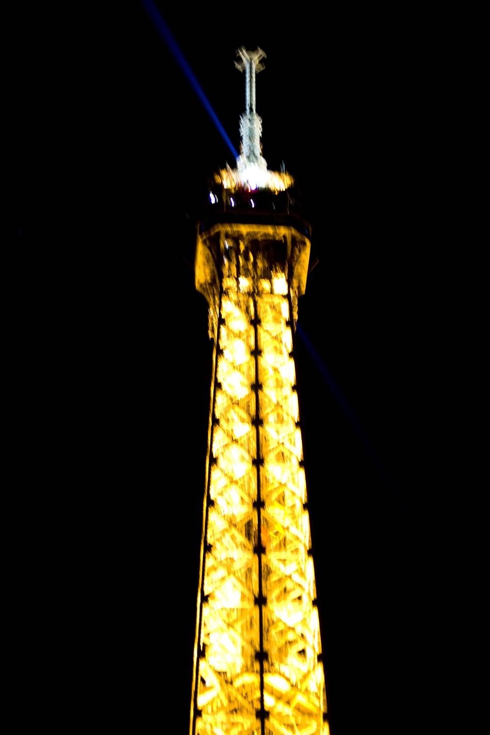 The lit spire and beacon at the top of the Eiffel Tower shine against the dark sky.