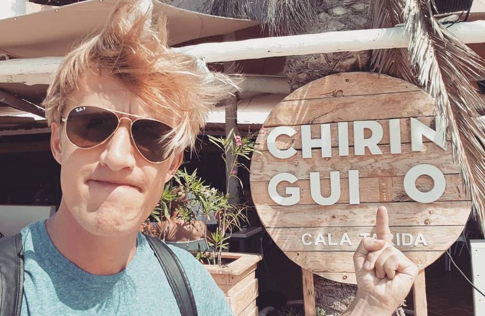 Selfie with windswept hair beside a wooden 'Chiringuito Cala Tarida' beach bar sign.