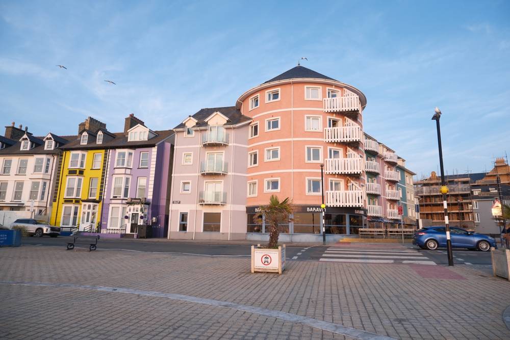 A picture of some multi-coloured buildings on a seafront. There's a small road diving them from where the photographer is stood.
a seagull on a lamp post is photobombing.