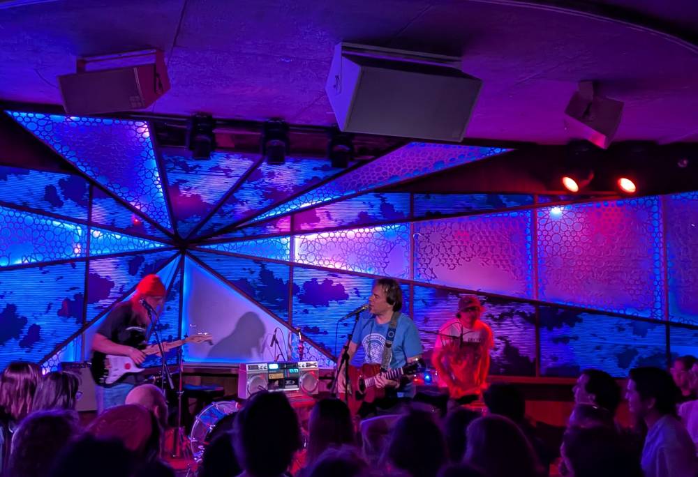 Emperor X band, guitar, base, and drums. Backlit by angular blue and purple lights.