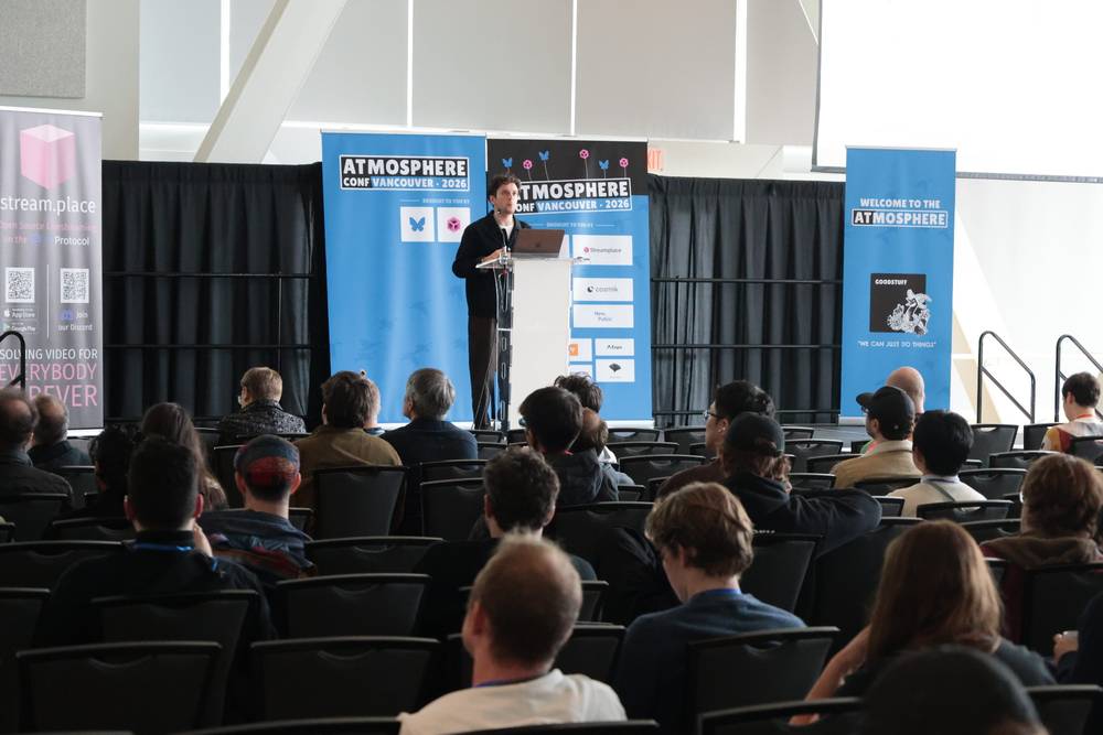 A man speaks at a podium on a conference stage in front of a seated audience.