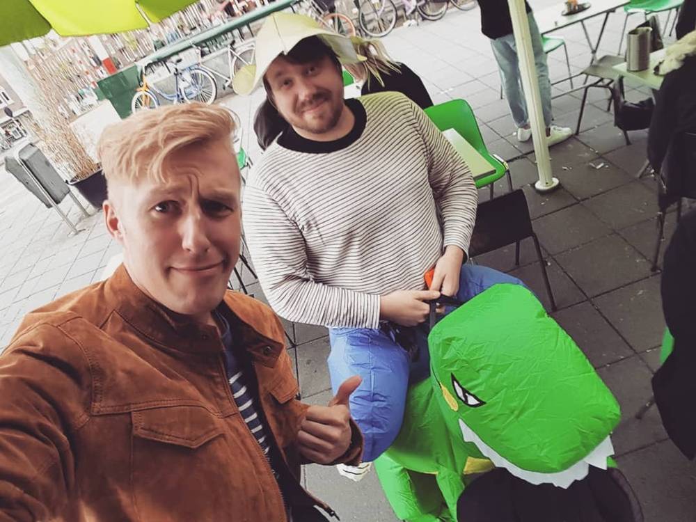 Selfie with a man and a large inflatable green dinosaur costume at an outdoor market.