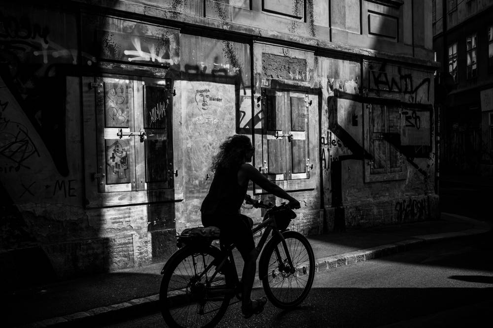 ⁠A low-angle black and white photograph of a figure riding a bicycle on a city street. The cyclist is seen in dramatic silhouette, backlit by a strong, low light source that creates a powerful halo effect and a very long, dark shadow cast directly towards the viewer. The background wall is heavily textured and marked with extensive graffiti and tags, featuring a closed wooden door with metal hinges. Deep pockets of impenetrable shadow dominate the scene, creating high contrast. The image is presented with a white border.