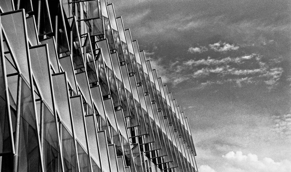 Close, oblique view of a glass-and-steel building facade. Dozens of triangular glass blades cantilever outward from a curtain wall in a regular rhythm, receding diagonally toward the upper left. Reflected clouds and structure ripple across their angled surfaces. To the right, a bright sky with scattered cumulus clouds fills the upper half of the frame.