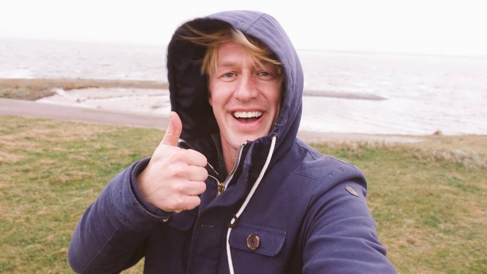 A man in a hooded jacket grins and gives a thumbs up on a windswept beach.