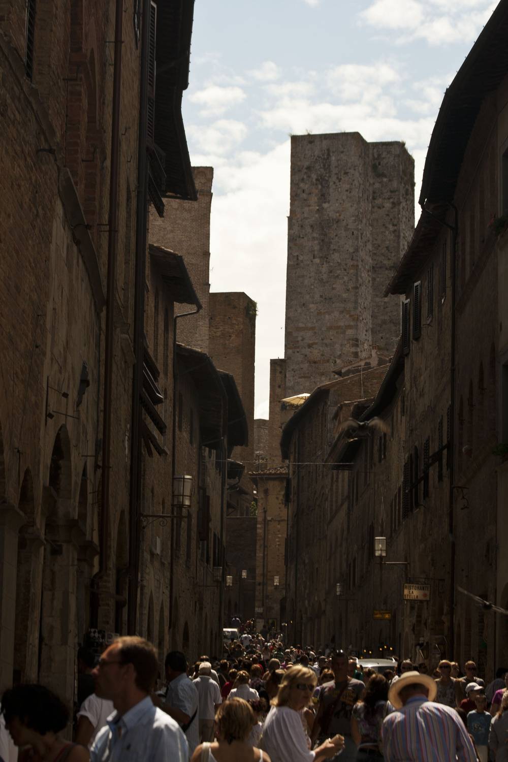 A crowd fills a narrow medieval street between tall stone towers.