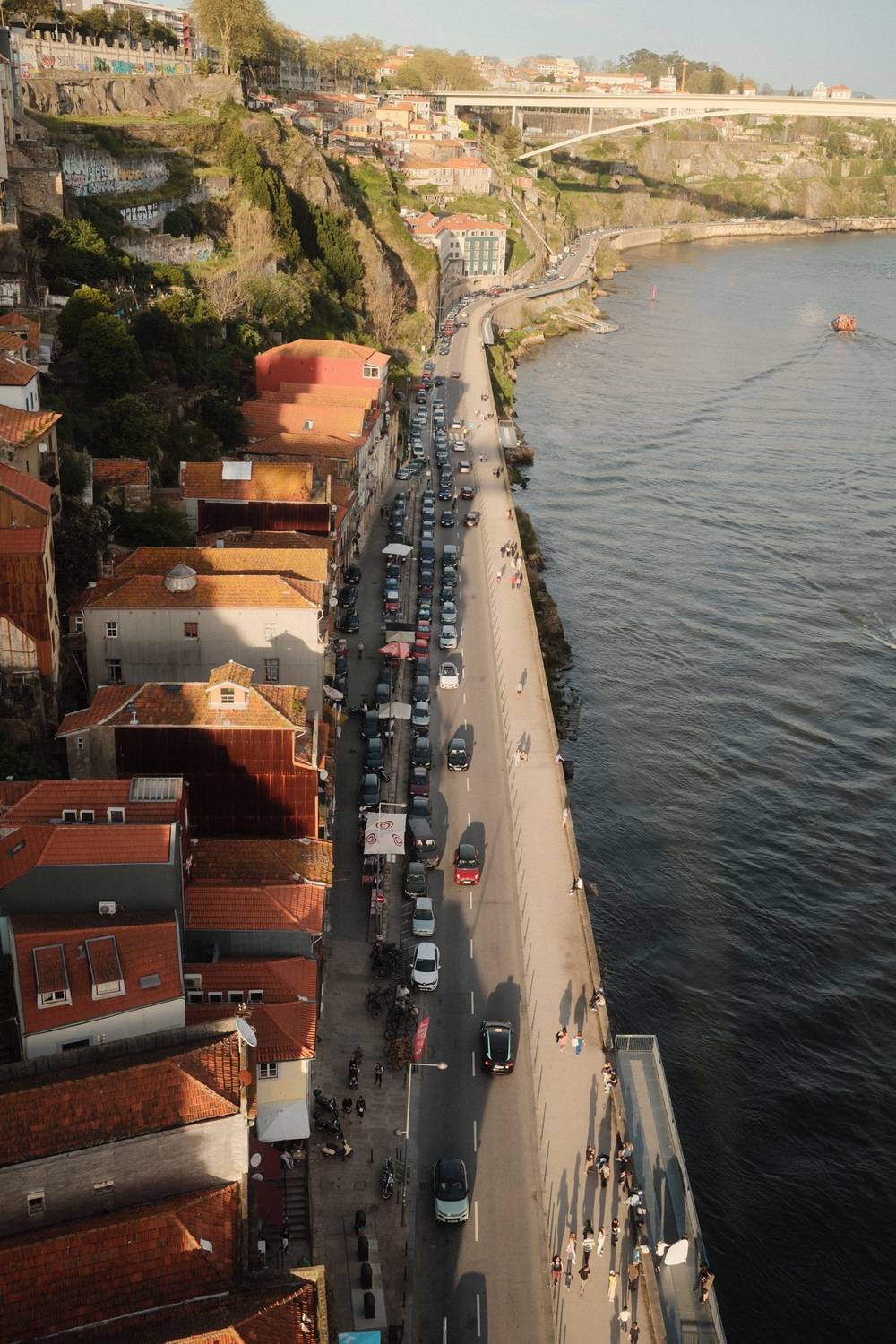 Bird's-eye view of Porto's Ribeira waterfront, showing a traffic-jammed riverside road between traditional red-roofed buildings and the Douro River in golden-hour light.