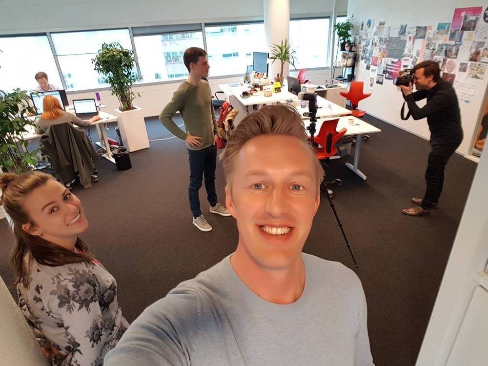 A man and woman smile for a selfie in an open-plan office while a colleague photographs them.