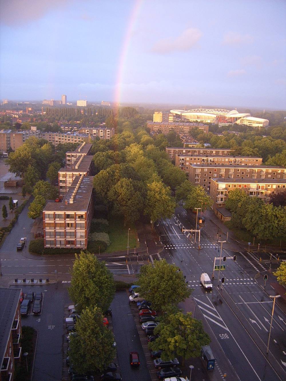 A rainbow arcs over a tower block and a stadium in Utrecht under a partly cloudy sky.
