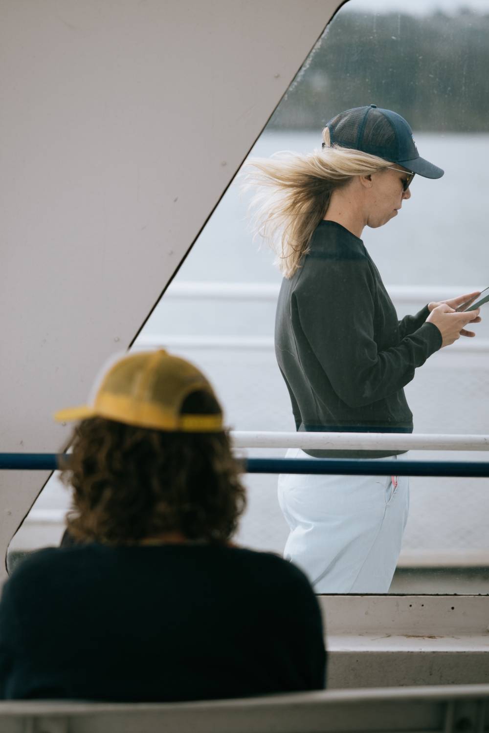 A woman with long blonde hair blowing in the wind walks along the deck of a BC Ferries vessel, looking at her phone. In the foreground, the back of another passenger wearing a yellow cap is visible. The ocean is visible through the ferry's windows behind her.
